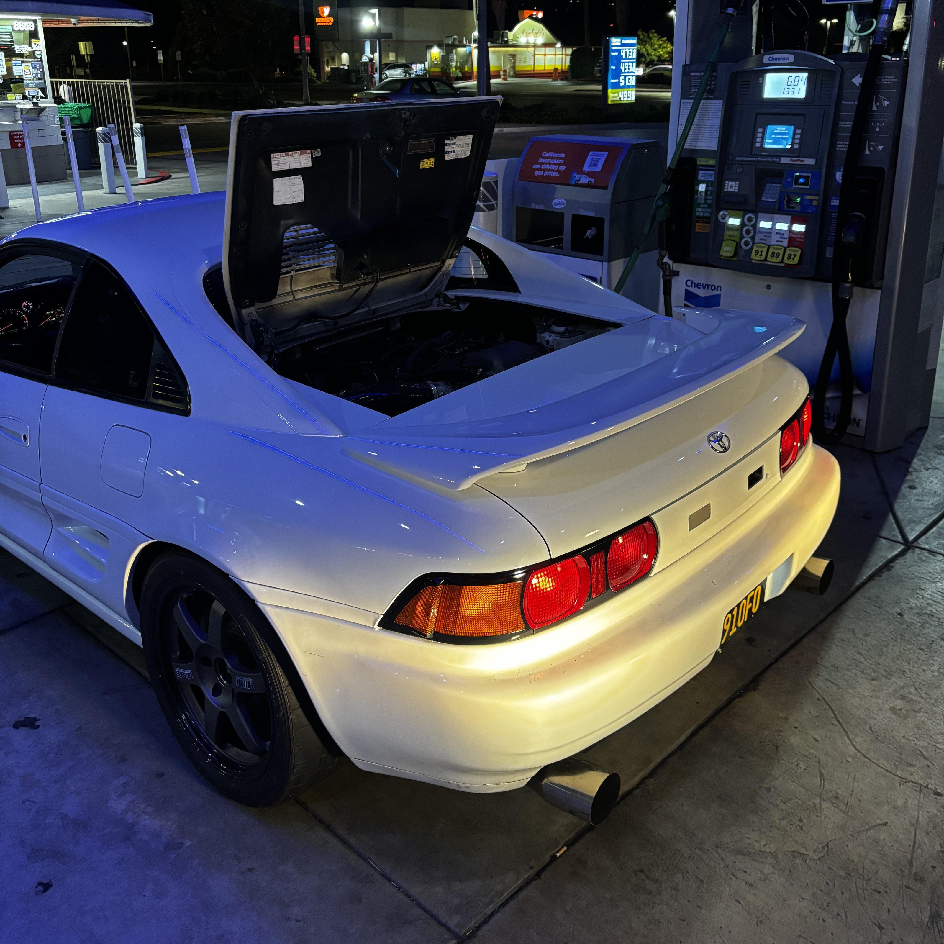 A white coupe parked at a gas station pump at night, with its trunk open and a fuel canister visible near the rear hatch. The car is lit by the station lights, and a Textron-branded fuel pump is visible beside it.