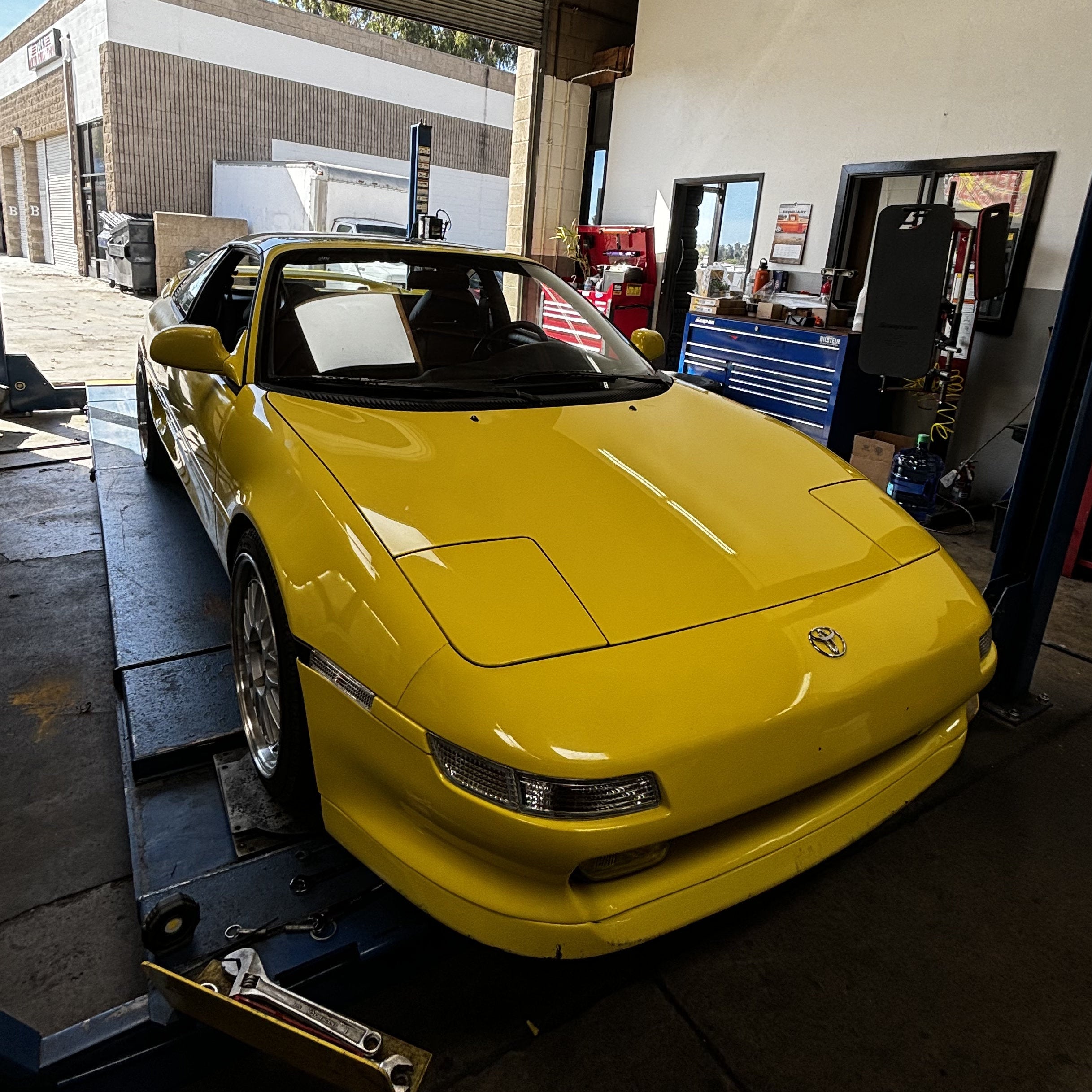 A bright yellow classic Toyota MR2 sports car is parked on a hydraulic lift inside a garage workshop.