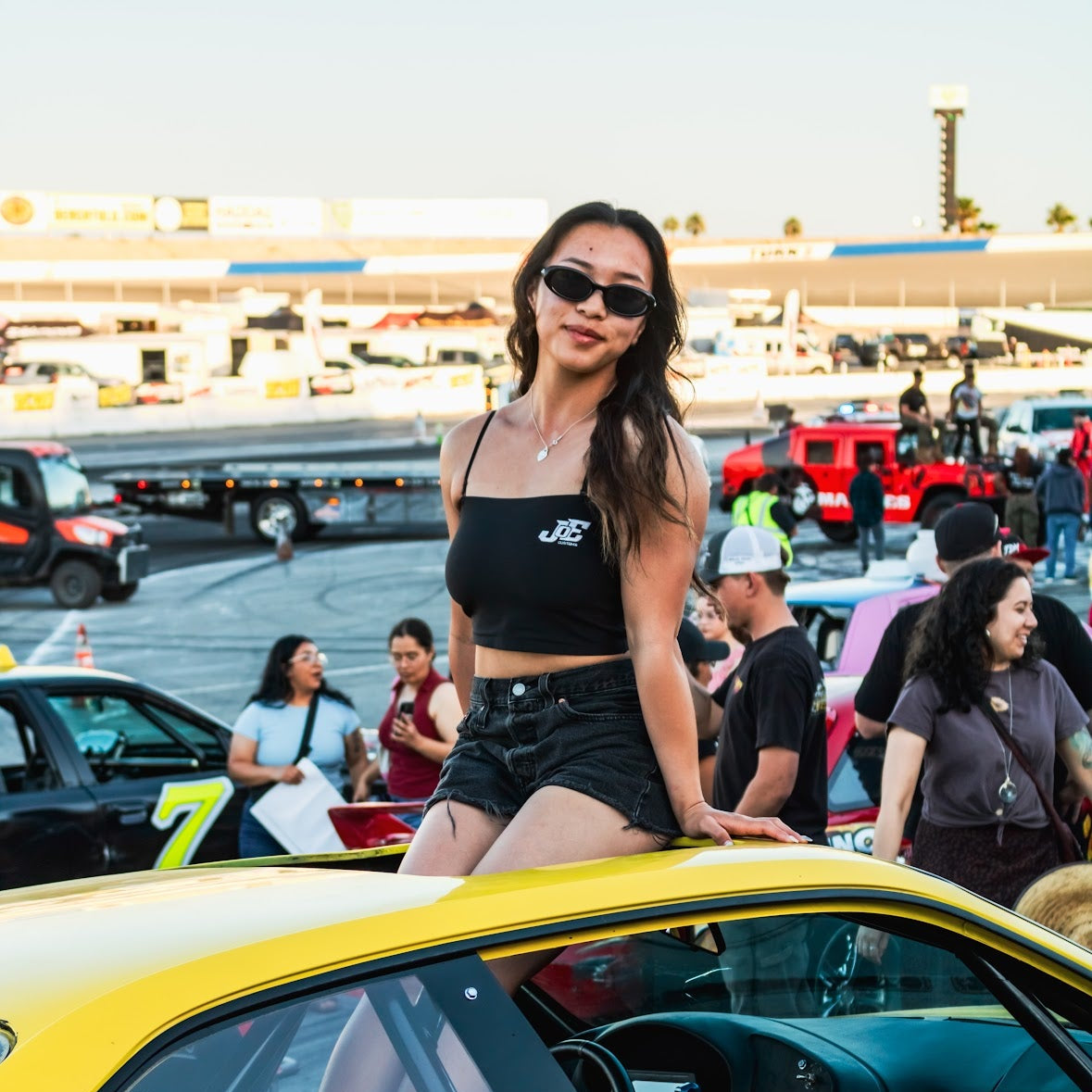 Woman posing on a yellow car with 'Joe Custom' branding at an outdoor event.